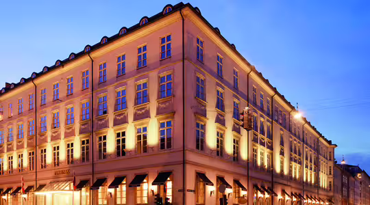 En stor, elegant bygning med varme lys, der oplyser facaden i skumringen. Det har en klassisk arkitektonisk stil med flere vinduer og en hjørneindgang. Himlen er klar, og gaden foran er tom.