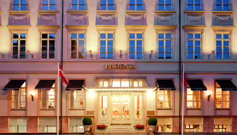 Billedet viser facaden på Hotel Phoenix med dets beige ydre og ensartede vinduer. "PHOENIX" vises tydeligt over indgangen, som er flankeret af potteplanter og to flag. Bygningen er varmt oplyst.