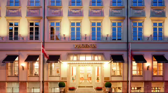 Billedet viser facaden på Hotel Phoenix med dets beige ydre og ensartede vinduer. "PHOENIX" vises tydeligt over indgangen, som er flankeret af potteplanter og to flag. Bygningen er varmt oplyst.