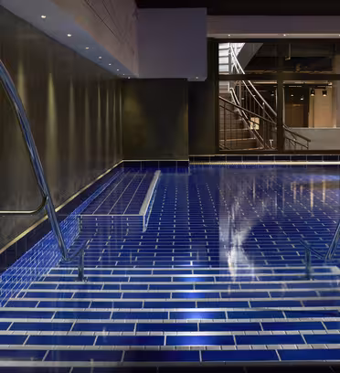 Indendørs pool med mørkeblå flisebelagte trin, der fører ud i vandet. Rustfrit stål håndlister på begge sider af trinene. Poolområdet er svagt oplyst med ovenlys og en vinduesvæg i baggrunden.