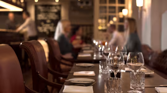 Svagt oplyst, elegant restaurant med et langbord dækket med vinglas, tallerkener og servietter. Læderstole står langs bordet, og slørede gæster sidder i baggrunden, hvilket skaber en hyggelig, intim atmosfære.