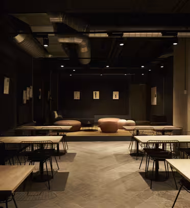En svagt oplyst moderne cafe med træborde og wireframe stole. Blød belysning fremhæver lyserøde og brune sofaer på en hævet platform. Industrielle elementer som synlige kanalsystemer og betonvægge bidrager til den moderne stemning.