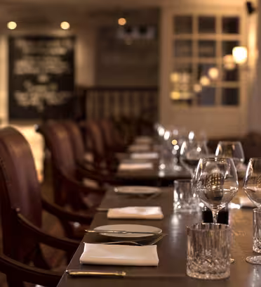 Et svagt oplyst, elegant restaurantinteriør med et langt bord til spisning. Bordet er beklædt med røde læderstole og pænt arrangeret glas, sølvtøj og hvide servietter. En tavle og blød belysning er synlige i baggrunden.