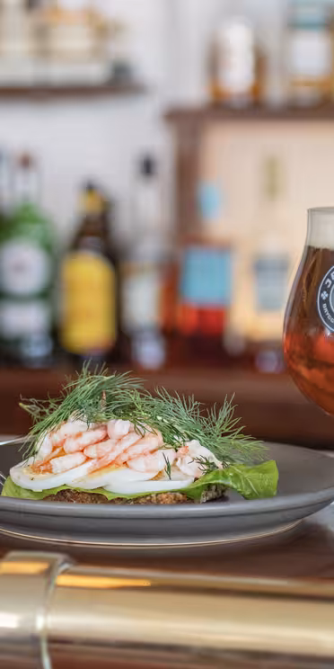 En tallerken med rejer og æg på salat pyntet med dild ved siden af et glas rav øl på en bardisk. Flasker og en bartender er sløret i baggrunden.