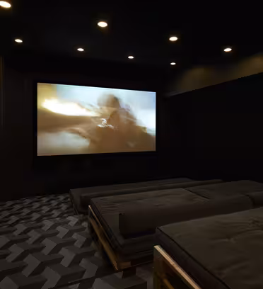Et svagt oplyst hjemmebiografrum med en stor skærm, der viser en sløret actionscene. Gulvet har et geometrisk mønster, og siddepladserne består af mørkegrå polstrede platforme. Forsænkede loftslamper tilføjer stemning til rummet.
