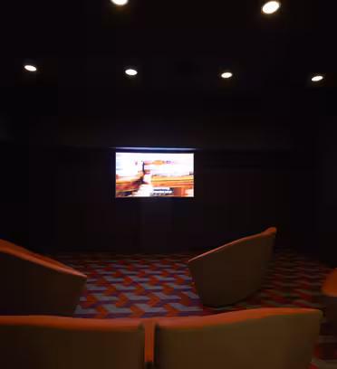 Et svagt oplyst rum med en stor skærm, der viser et billede på den fjerneste væg. Fire moderne, orange stole er arrangeret foran skærmen. Gulvet er dækket af et geometrisk mønstret tæppe.