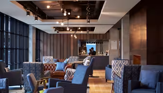 Moderne lounge med blå og grå lænestole, en brun lædersofa og hængende lysarmaturer. Et barområde med en person, der arbejder, er synligt i baggrunden. Rummet har store vinduespartier og trægulve.