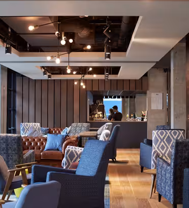 Moderne lounge med blå og grå lænestole, en brun lædersofa og hængende lysarmaturer. Et barområde med en person, der arbejder, er synligt i baggrunden. Rummet har store vinduespartier og trægulve.