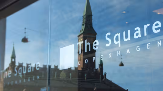 Refleksion af en gammel bygning med et grønt spir i glasvinduet på "The Square" hotel i København. Refleksionen viser indviklede arkitektoniske detaljer af bygningen under en klar himmel.