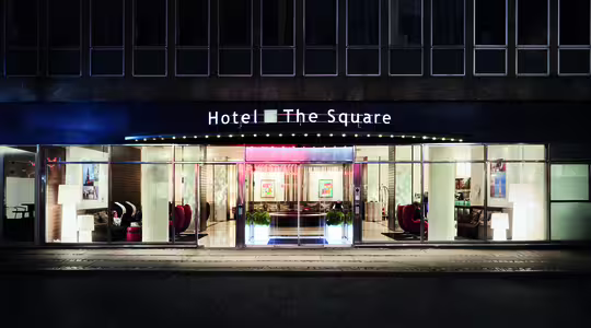Gadebillede af en hotelindgang ved navn "Hotel The Square" om natten. Bygningen har store vinduer, der viser en veloplyst lobby med moderne møbler, kunstværker og potteplanter. Det oplyste skilt tilføjer et indbydende touch.