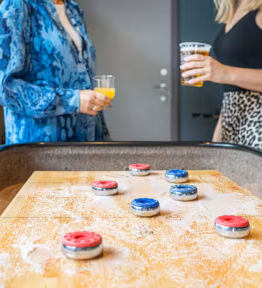 Nærbillede af et shuffleboard-spil med røde og blå pucke på en træoverflade. To personer står i baggrunden, den ene i en blåmønstret skjorte og holder en drink, og den anden i en sort top med en nederdel med leopardprint og holder også en drink.