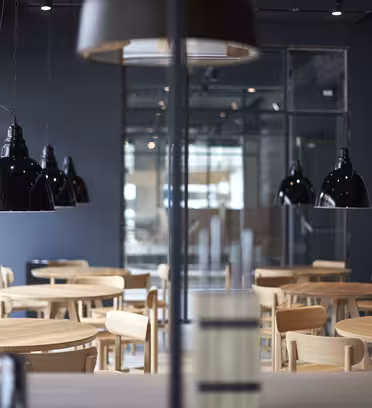 En moderne, tom spiseplads med runde træborde og stole. Sorte pendler hænger fra loftet, og store vinduer tillader naturligt lys at fylde rummet. Den overordnede indretning er minimalistisk og industriel.
