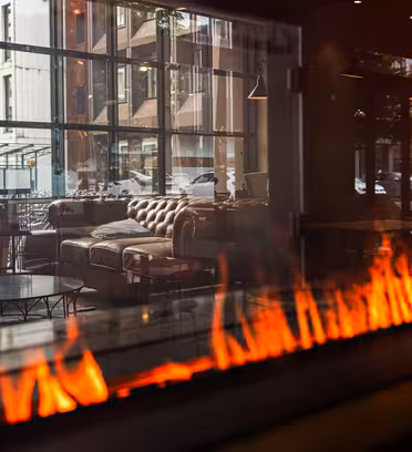 Et hyggeligt interiør med en brun lædersofa og et glas sofabord ses gennem et vindue. En moderne pejs med flammer tilføjer varme til omgivelserne. Refleksionen viser et bybillede med kontorbygninger i baggrunden.