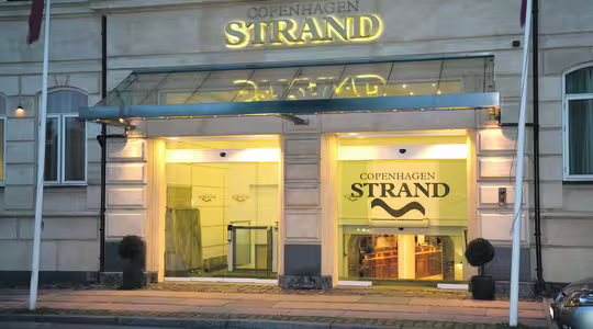 Indgang til Copenhagen Strand Hotel i skumringen, med en glasmarkise med hotellets navn oplyst i guld. Facaden er lysfarvet med to små potteplanter, der flankerer indgangen.