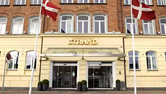Facade på Copenhagen Strand Hotel med glasindgang, flankeret af to danske flag på stænger. Murstensbygningen har året "1869" vist ovenfor og flere vinduer med hvide rammer.
