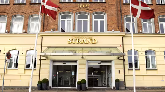 Facade på Copenhagen Strand Hotel med glasindgang, flankeret af to danske flag på stænger. Murstensbygningen har året "1869" vist ovenfor og flere vinduer med hvide rammer.