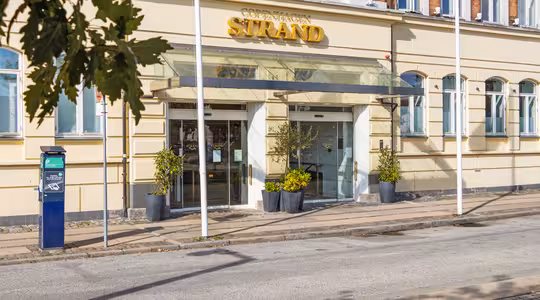 Indgang til Copenhagen Strand Hotel, med en beige facade, to potteplanter, en baldakin og en synlig parkeringsautomat på en solskinsdag.