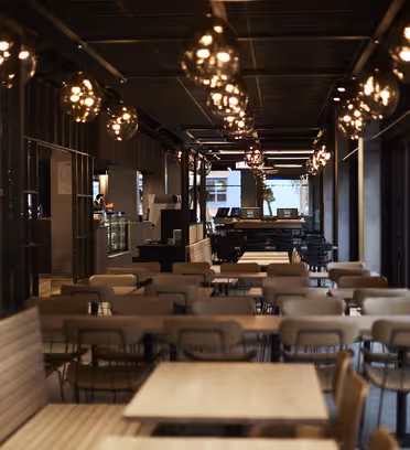 En moderne, svagt oplyst cafe med rækker af tomme træborde og stole. Varme pendler hænger fra loftet og skaber en hyggelig atmosfære. En tæller er synlig i baggrunden.