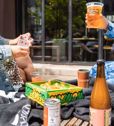 Folk spiller kort ved et picnicbord med drinks og en kasse chips. To personer, der holder drikkevarer, ses, klædt i afslappet, farverigt tøj. En ølflaske og en dåsedrik står på bordet med et mørkt tæppe under.