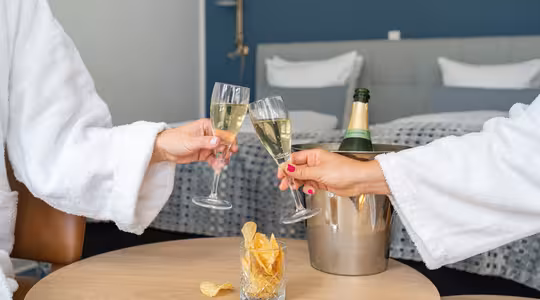 To personer i hvide rober skåler med champagneglas på et stilfuldt hotelværelse. En flaske champagne hviler i en isspand på bordet sammen med et glas fyldt med kartoffelchips. Plys sengetøj er synligt i baggrunden.