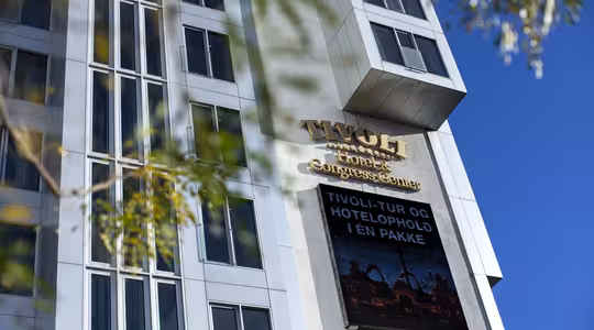 Høj moderne bygning med et skilt med teksten "Tivoli Hotel & Congress Center" med en reklameskærm nedenfor. Bygningen har lodrette vinduer og er sat mod en klar blå himmel med trægrene delvist i rammen.