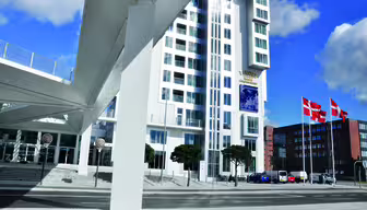 Høj moderne bygning med hvid facade og geometriske designdetaljer, med danske flag på stænger foran. En fodgængerbro krydser over gaden, der forbinder bygningen. Et par biler er parkeret nær indgangen.