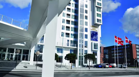 Høj moderne bygning med hvid facade og geometriske designdetaljer, med danske flag på stænger foran. En fodgængerbro krydser over gaden, der forbinder bygningen. Et par biler er parkeret nær indgangen.