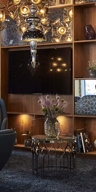 En moderne stue med stilfulde møbler, herunder to blå lænestole og en sofa omkring et glas sofabord. En træreol med dekorative genstande og et tv er i baggrunden. Dekorativ belysning tilføjer varme til scenen.
