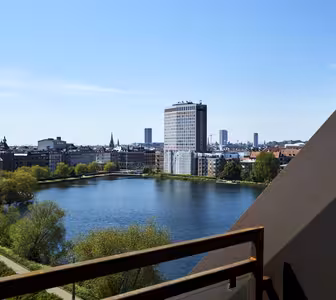 Udsigt over et bybillede med en sø med træer langs kanterne, en høj moderne bygning med en synlig vandfaldsfacade og flere andre bygninger under en klar blå himmel, set fra en balkon.