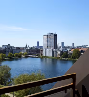 Udsigt over et bybillede med en sø med træer langs kanterne, en høj moderne bygning med en synlig vandfaldsfacade og flere andre bygninger under en klar blå himmel, set fra en balkon.
