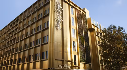 En moderne bygning i flere etager med udvendig guld og lodret havemur. Designet har store vinduer og et skilt, hvor der står "Nhow Hotel." Træer med efterårsblade er i forgrunden under en klar blå himmel.