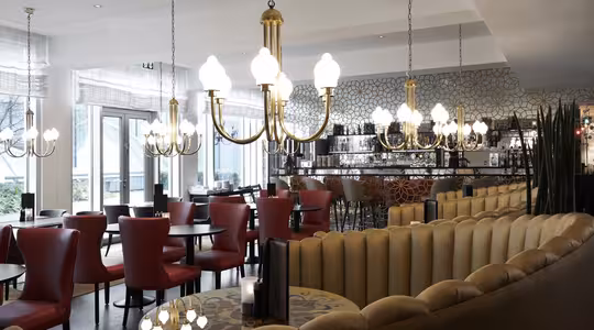 En rummelig restaurant med elegant belysning har lysekroner, der hænger fra loftet. Siddepladserne inkluderer røde stole og beige polstrede kabiner. En mønstret væg og en bar med høje taburetter ses i baggrunden. Store vinduespartier bringer naturligt lys ind.