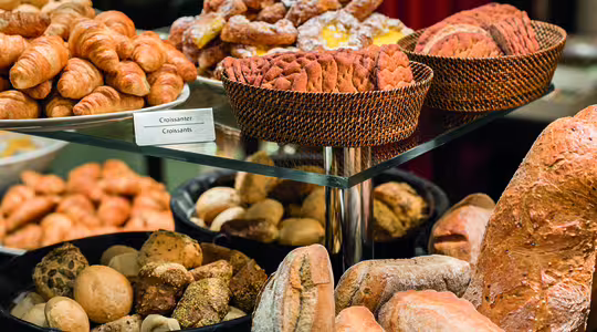 Diverse kager og brød vist på en glashylde, inklusive croissanter, kager i pulverform og forskellige brød. De er arrangeret i flettede kurve og runde bakker, hvilket skaber en tiltalende og mangfoldig bagerpræsentation.