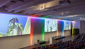 Et auditorium med en moderne opsætning byder på farverig LED-belysning langs væggene. Et langt bord med stole er placeret foran, og store skærme viser arkitektoniske billeder. Siddeområdet består af flerfarvede stole.
